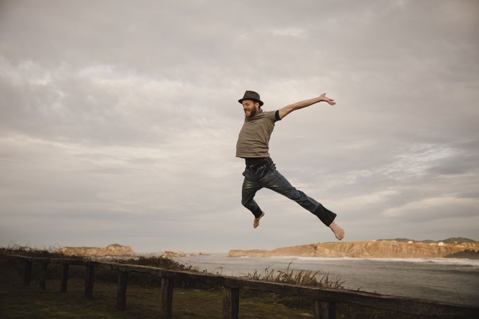 Young man in hats having fun on seat on coast near waving sea and cloudy sky