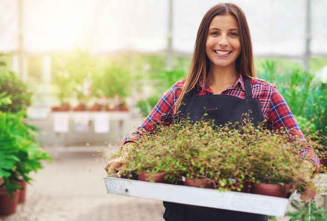 Smiling woman holding tray of plants for sale in a greenhouse