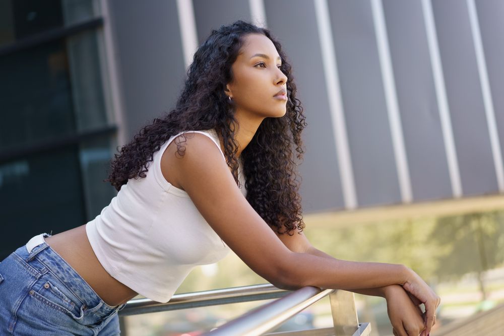 A thoughtful young woman is leaning against a railing in an urban setting during the day