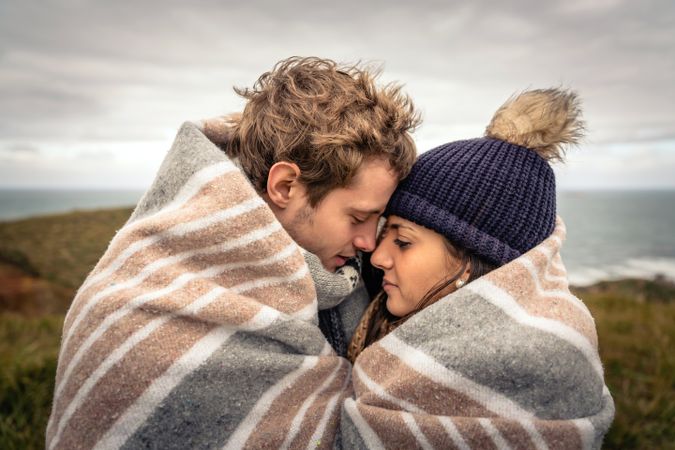 Closeup of young couple embracing under blanket in a cold day with sea and dark cloudy sky in the background