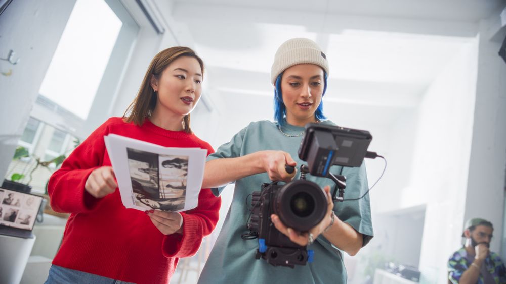 Two Women Engaged In A Film Production Session, One Holding A Camera And Other With Script, In A Bright, Modern Studio, Showcasing Teamwork In Creative Filmmaking And Advertising.