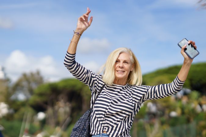 Older woman in jeans and striped shirt outside on sunny day with arms up holding smart phone