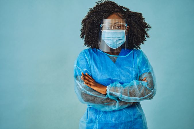 Black female medical professional in surgical PPE, eye mask and shield, with her arms crossed