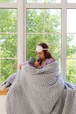 Girl sipping from mug, wrapped in knitted blanket by window.