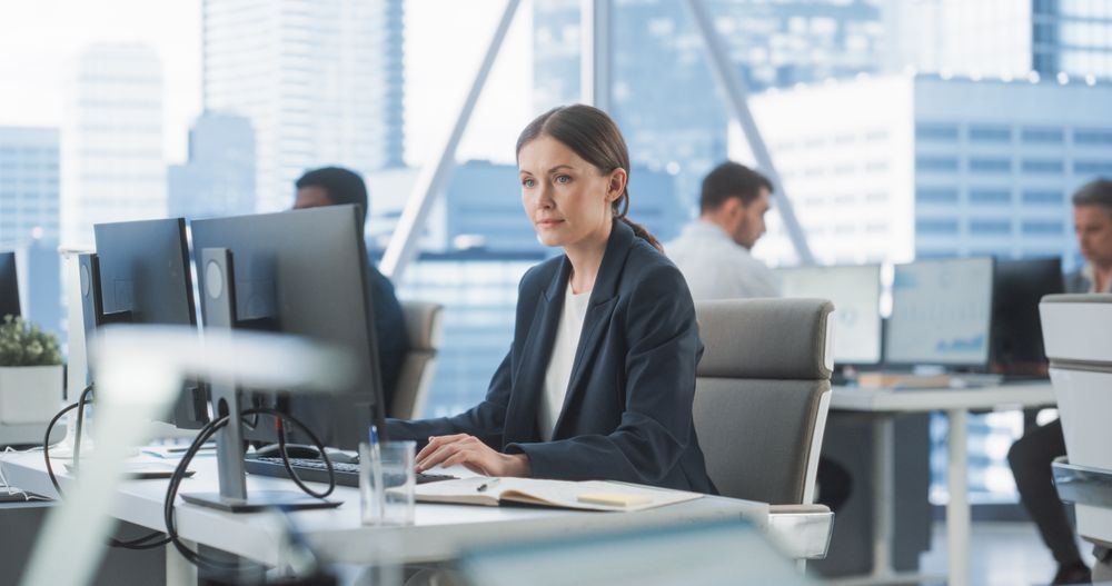 Focused Female Operations Specialist Typing On Desktop Computer In Diverse Corporate Office With Megapolis Window View.