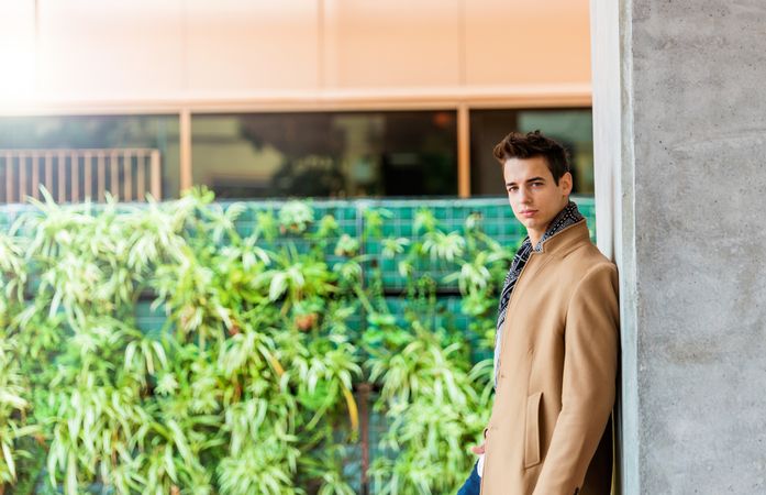 Side view of handsome man wearing camel coat and scarf looking at camera against cement