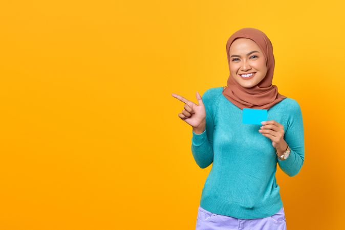 Muslim woman pointing to the side and holding credit card in yellow studio