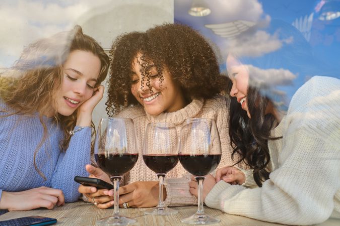 Smiling multiethnic women with wine looking at phone screen