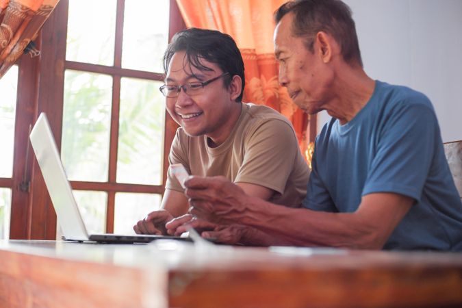 Happy adult son and mature father pay online using laptop and credit card