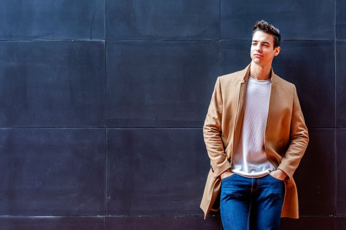 Cool male in camel coat looking up leaning against dark wall