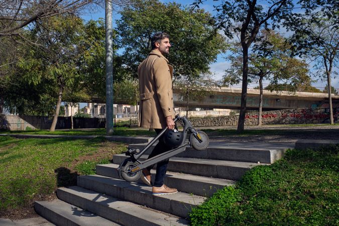 Stylish man with electric scooter walking up stairway in park