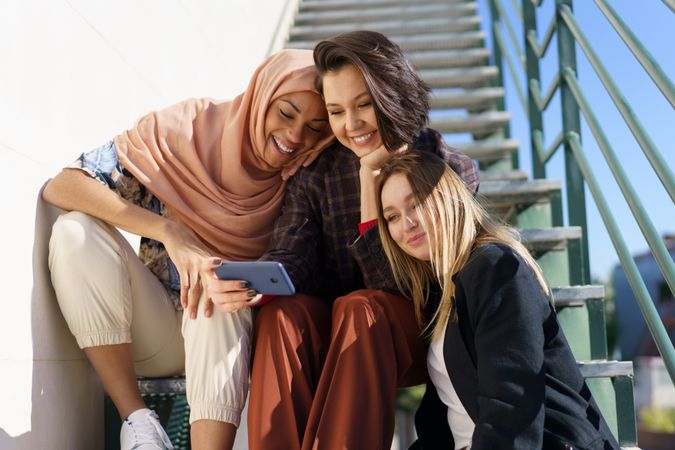 Multi-ethnic women friends in stylish outfits looking at phone on metal stairs outside