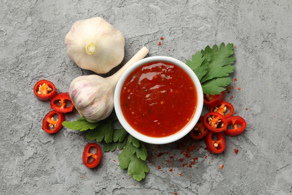 Bowl with chilli sauce, pepper and garlic on gray background
