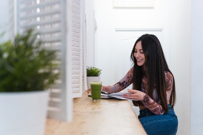 Delighted woman taking notes in notebook in bar with cocktail