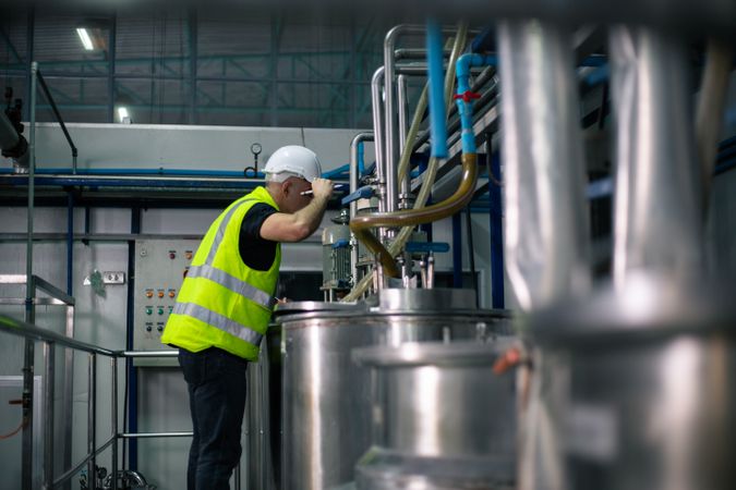 A male industrial engineer wearing a safety helmet and reflective vest is inspecting equipment in a factory. He is inspecting machine system. Background show industrial pipes and machinery
