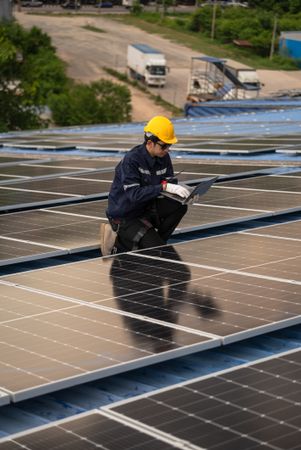 Male engineer checking solar cell or photovoltaic cell by computer laptop installed on the roof of the factory. Technician worker repair or inspect the system. Industrial renewable energy concept