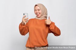 Happy Muslim woman in headscarf and orange shirt with phone and giving thumbs up 5oe7Gb