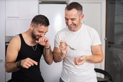 Laughing male couple tasting pasta as they prepare meal
