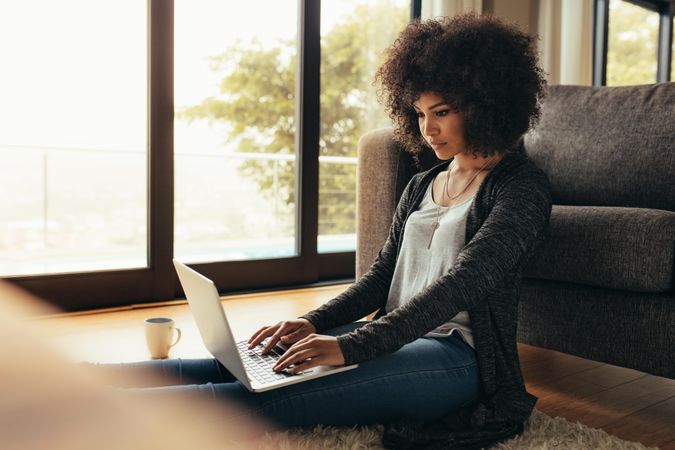 Young woman sitting on floor by the couch using laptop