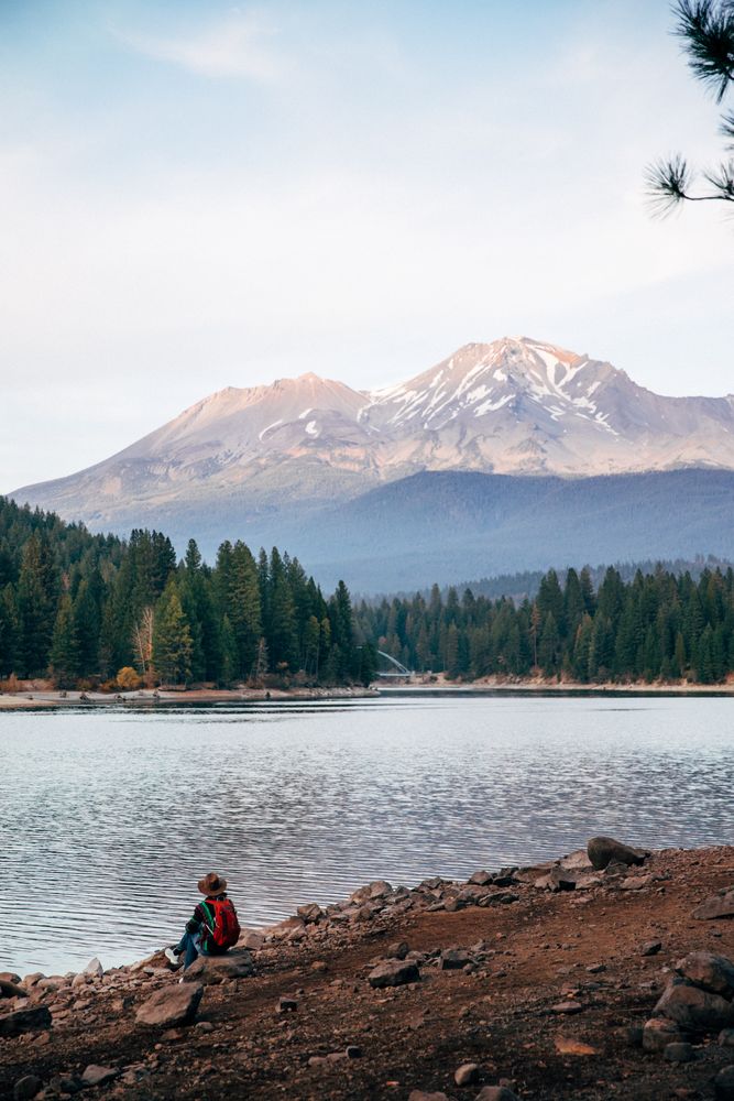 Person with fedora hat sitting on river shore in woods near Mt. Shasta in Siskiyou County, CA, US