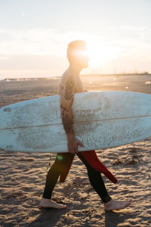 Tattooed man with surfboard under his arm at sunset