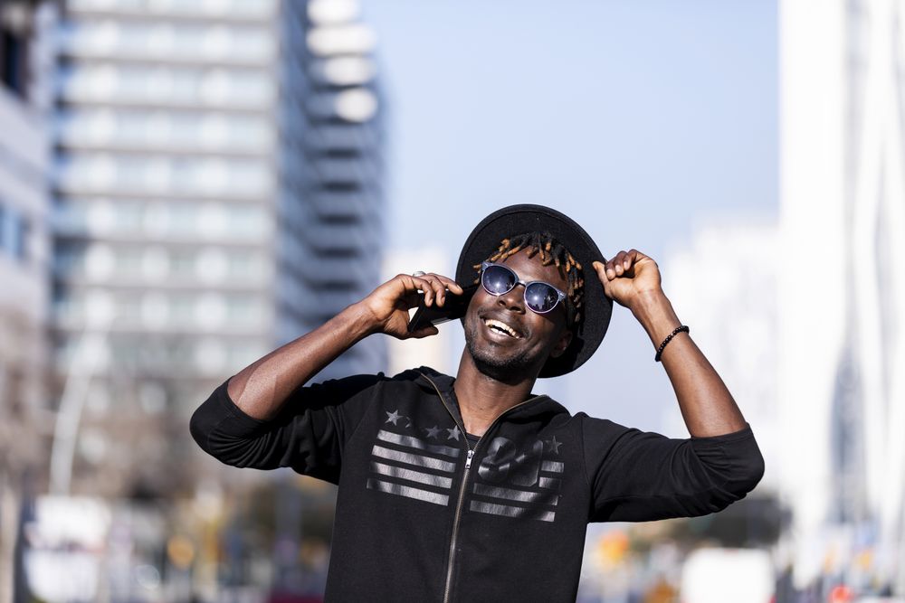 Happy Black man wearing hat & sunglasses standing on the street talking on cellphone