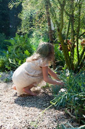 Profile of a young girl wearing a dress reaching for a plant on a rock path in garden