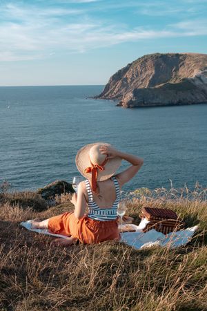 Woman with glass of drink on picnic mat looking at sea and mountains