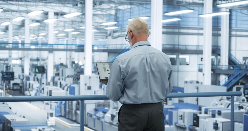 Portrait of an Adult Engineer Standing on a Platform, Using Laptop Computer at an Electronics Factory. Technician Making Technical Upgrades Online Over an Internet Software
