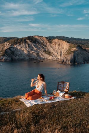 Woman with glass of drink on picnic mat looking at sea and mountains