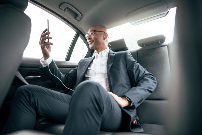 Businessman making a video call holding his cell phone while sitting in his sedan on back seat