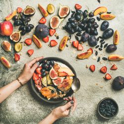 Woman holding bowl of yogurt with spoon over table of figs, peaches, and chia seeds, square crop