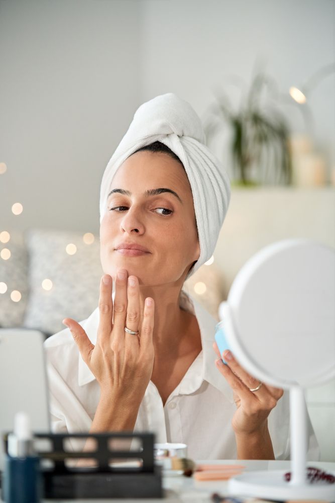 Attractive female in towel turban applying facial cream at mirro