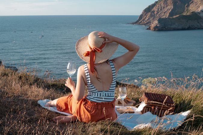 Woman with glass of drink on picnic mat looking at sea and mountains