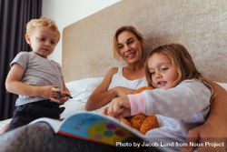 Cute little girl pointing at storybook while sitting with her mother and brother on bed 5oEO1b
