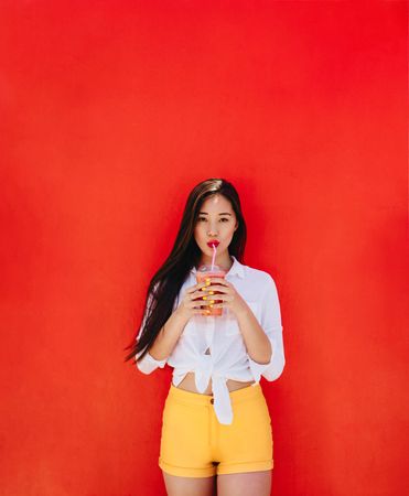 Young woman drinking juice through a straw and standing against red wall
