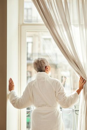 Woman with grey hair pulling back curtains of out tall window
