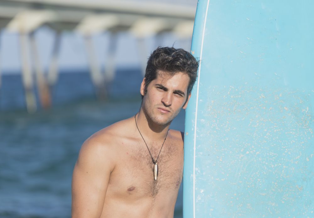 Front of shirtless male surfer standing next to blue board on the coast