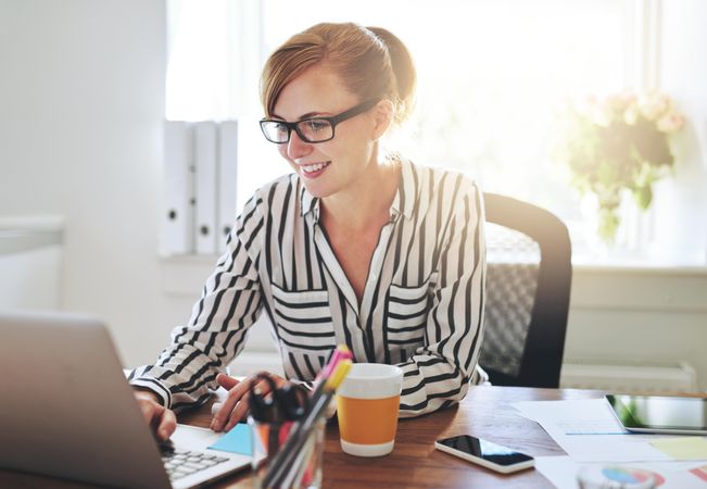 Happy woman working on laptop in bright home office with coffee