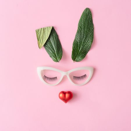 Bunny rabbit face made of natural green leaves with glasses and red heart on pastel pink background