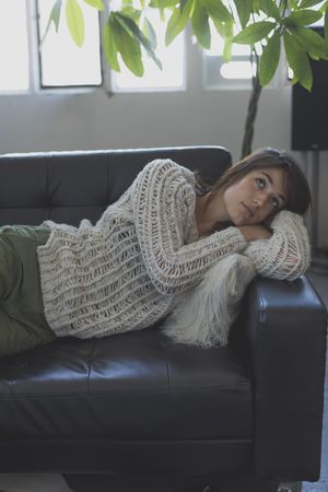Female in wooly sweater lying on her side trying to nap on leather couch in living room, vertical
