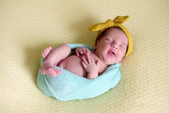 Close-up of a newborn baby girl, inside a ball of blue wool on a yellow blanket with a yellow bow on her head