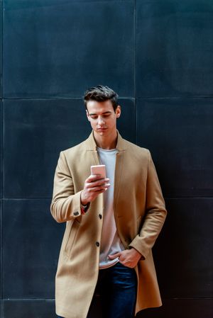 Serious man in fall coat leaning back on dark outdoor wall while texting