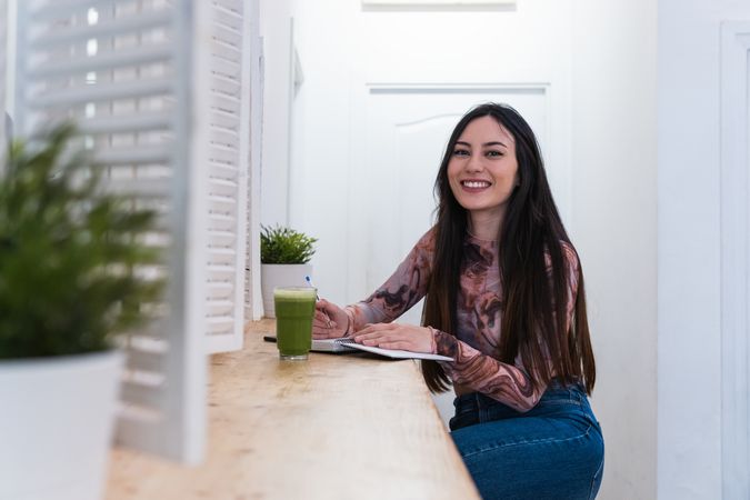Delighted woman taking notes in notebook in bar with cocktail