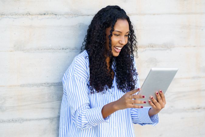 A happy and young woman is fully engaged while using her tablet against a modern wall