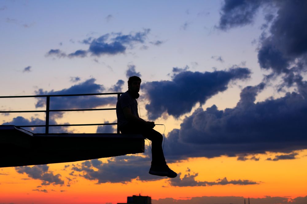 Out line of male sitting on ledge with phone against a colorful dusk side, vertical