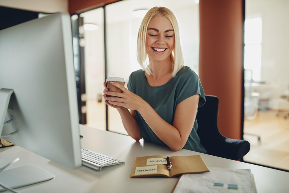 Blonde businesswoman smiling and holding a coffee at her work computer