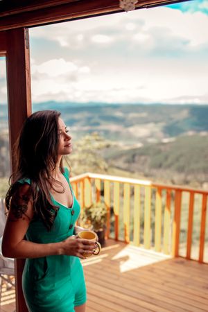 Woman wearing teal outfit holding a brown mug leaning on terrace door