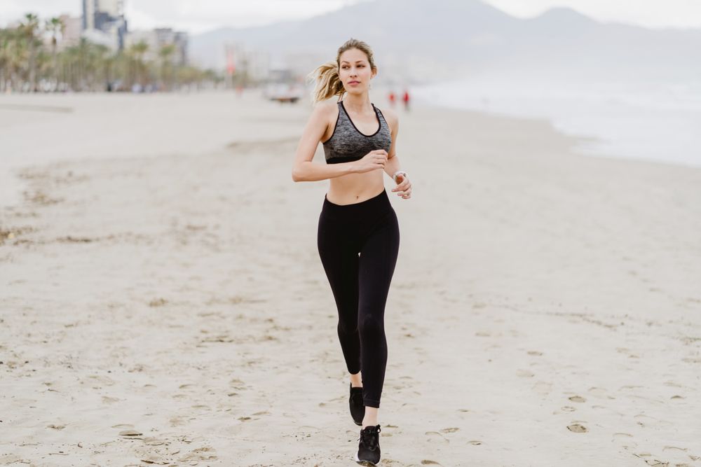 Female runner in stylish sportswear jogging along calm sandy seaside