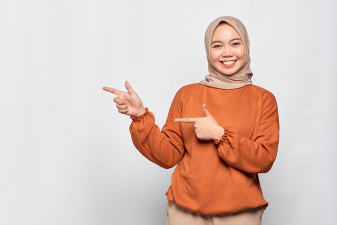Upbeat Muslim woman in headscarf and orange sweater pointing to her side